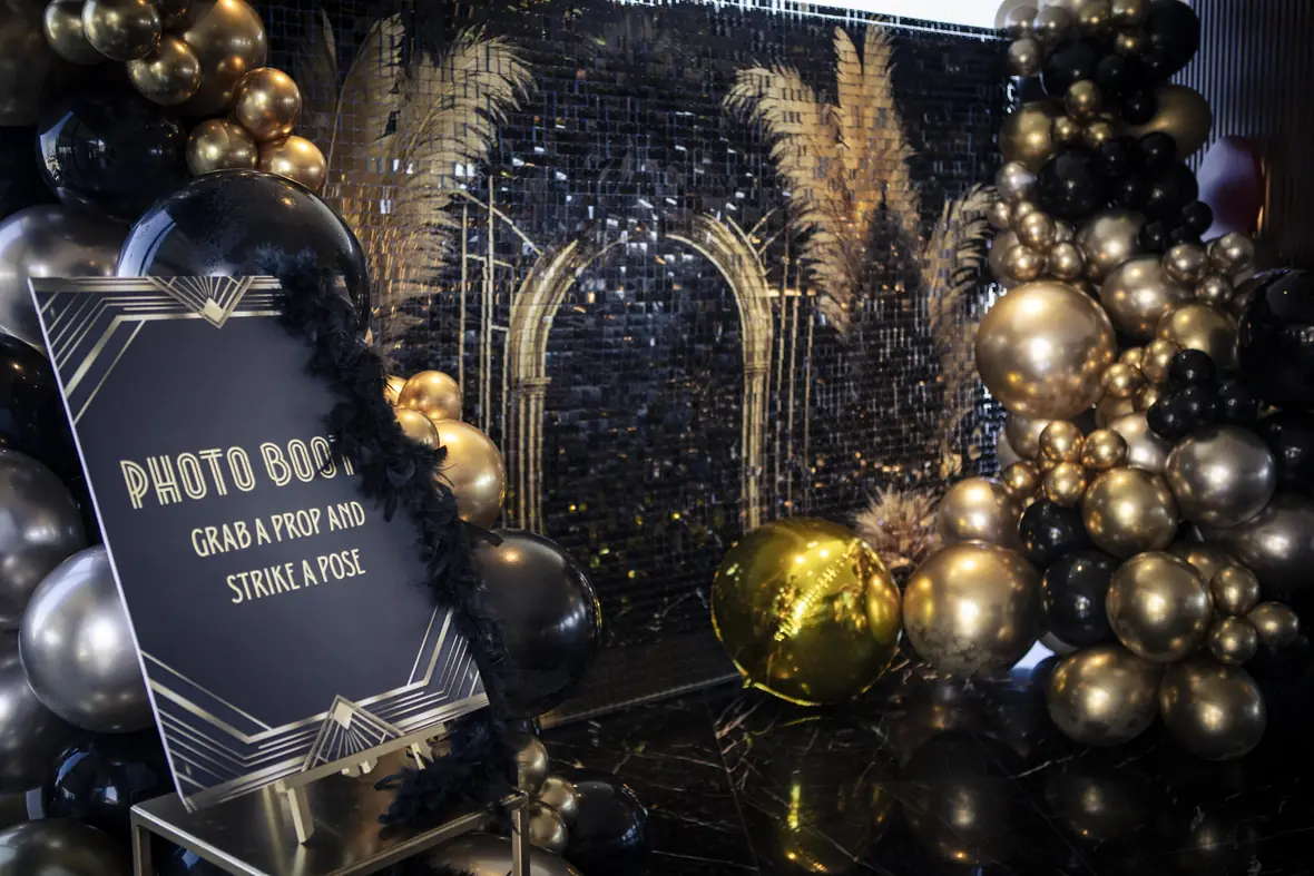 Black and gold photo booth backdrop with pampas grass and balloon garlands