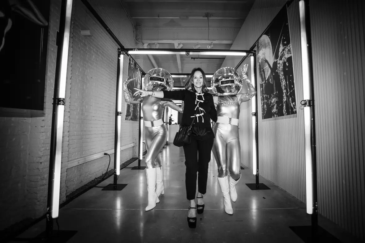 Guests walking a red-carpet corridor framed by columns of neon light, flanked by mirror-ball performers, in black and white