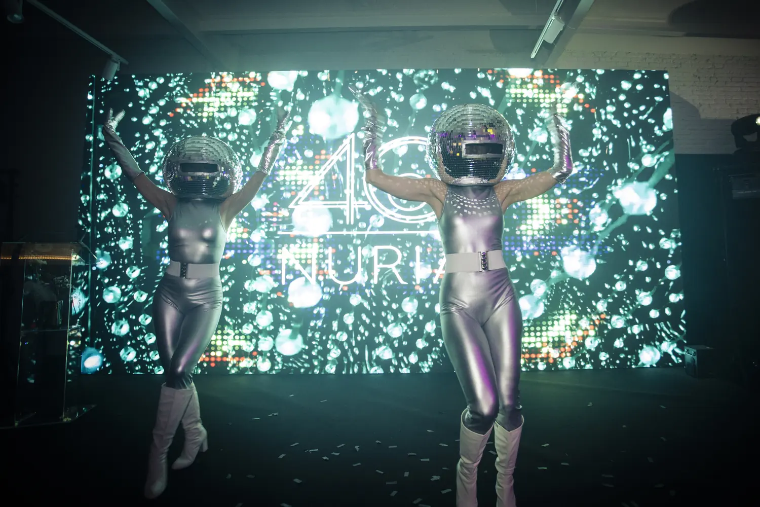Two performers in silver catsuits and mirror-ball helmets before a turquoise wall of light at a private 40th birthday