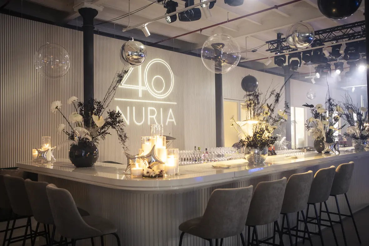 White-panelled club interior with an illuminated 40 Nuria monogram, dark florals, mirrored spheres and a candlelit bar