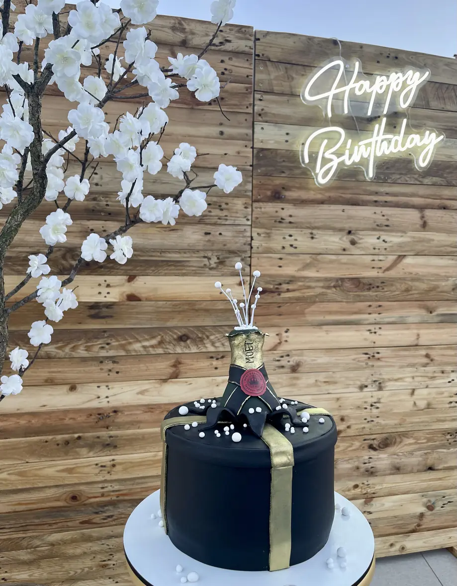 Black birthday cake with gold ribbon in front of a blossom and neon backdrop