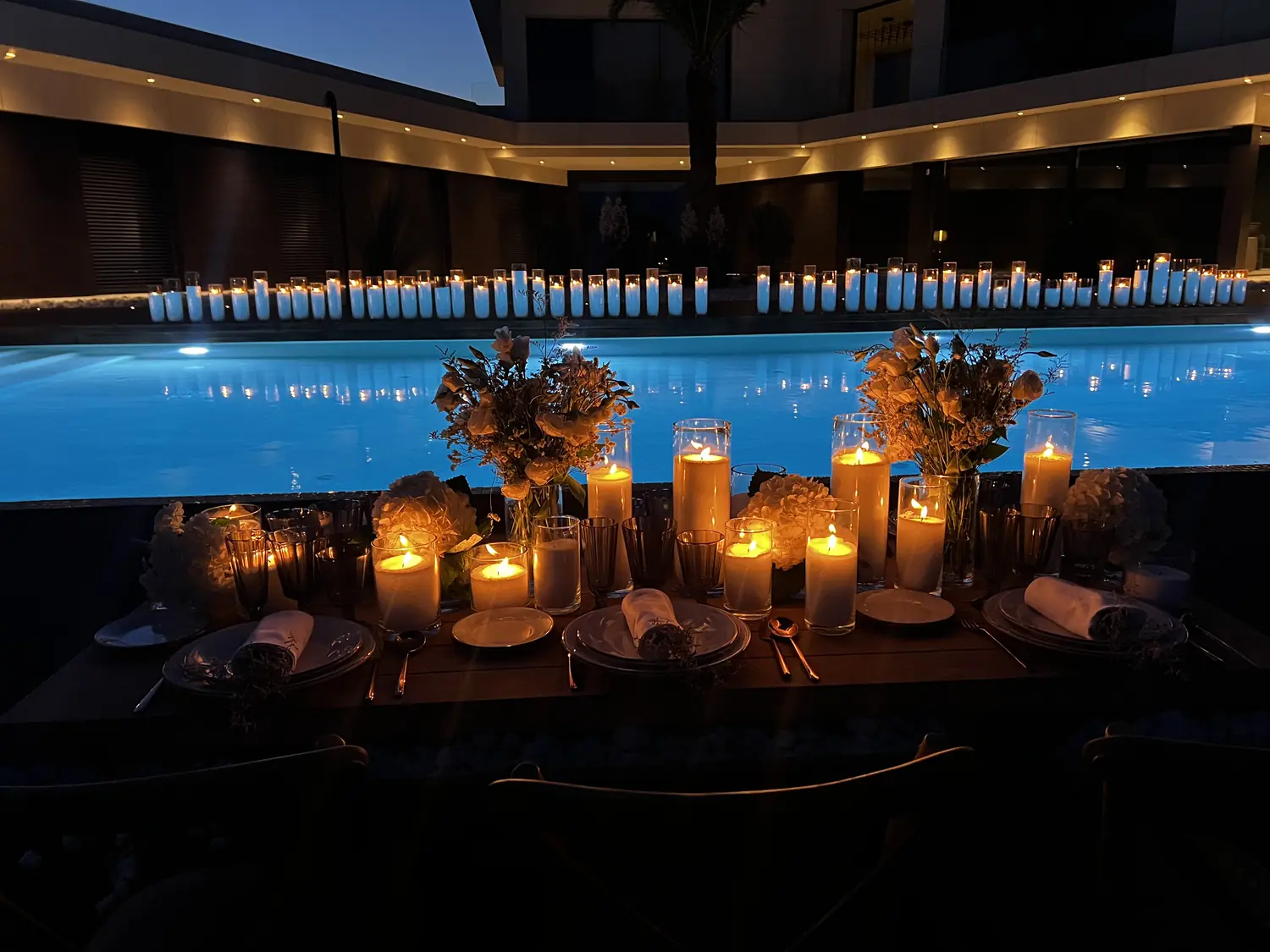 Candlelit dinner table beside a glowing blue pool at night, rows of pillar candles reflected in the water